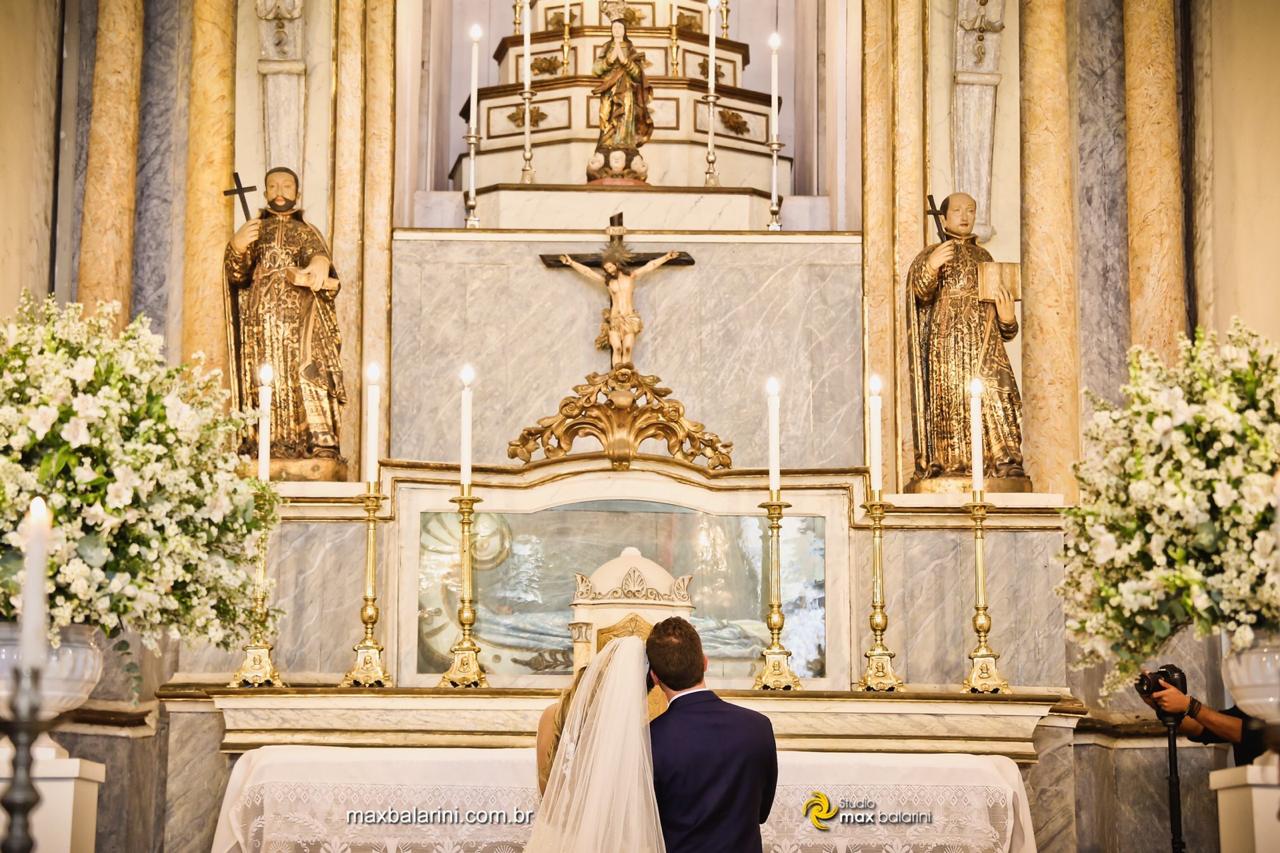 Casal de noivos de costas ajoelhados no altar ornamentado em ouro da igreja durante a benção nupcial.