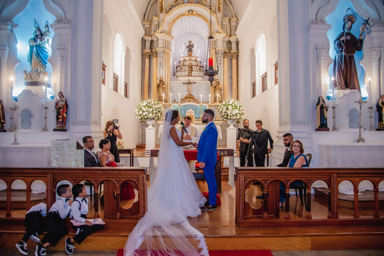 Noivos de mãos dadas durante os votos matrimoniais em altar ricamente adornado com imagens sacras e flores brancas.