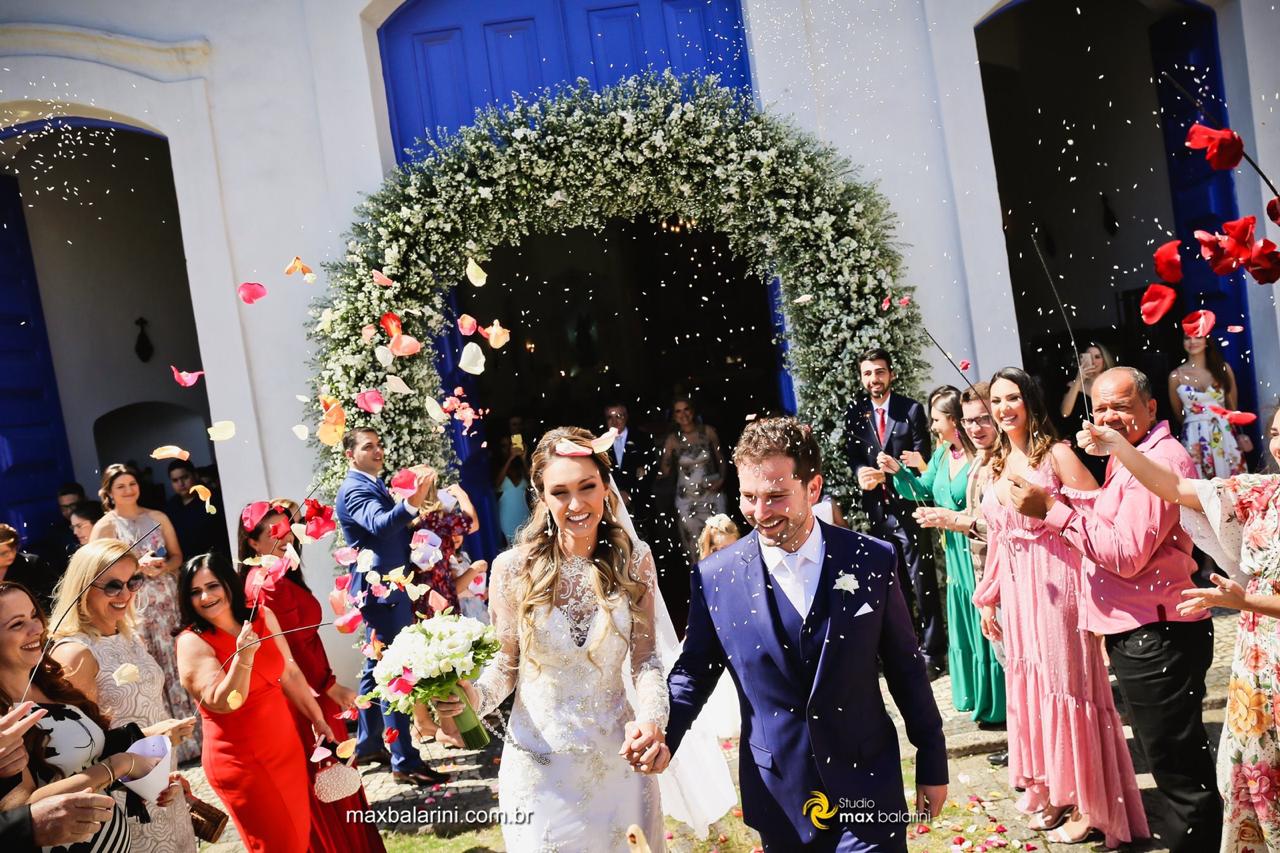 Noivos saindo da igreja sob arco de flores e chuva de pétalas de rosas celebrada pelos convidados.