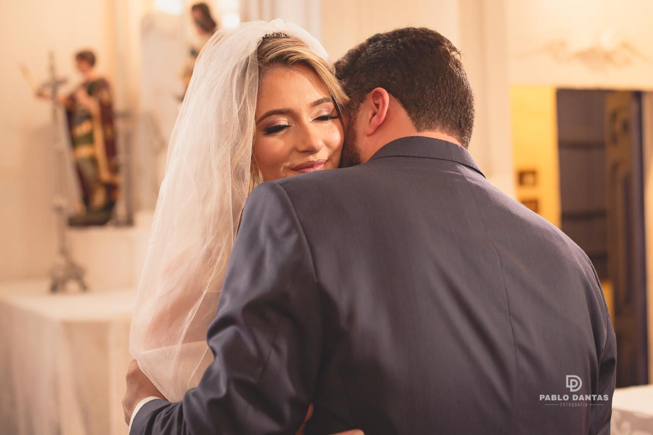 Close emocionante do noivo abraçando a noiva, que sorri de olhos fechados com seu véu em destaque.