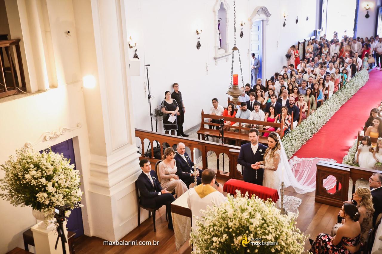 Foto tirada de um ângulo elevado mostrando os noivos no altar, convidados nos bancos e a decoração clássica da igreja.