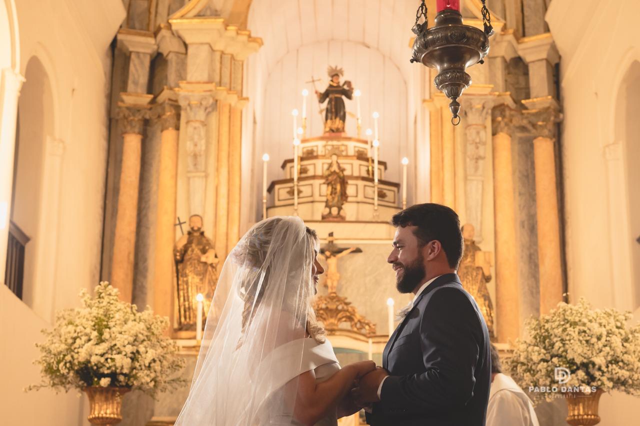 Noivos sorrindo um para o outro em frente ao altar histórico iluminado por velas e decorado com flores brancas.