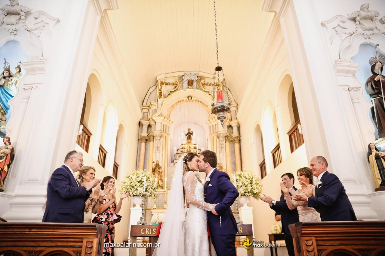 Noivos se beijando no altar sob aplausos dos padrinhos e madrinhas.