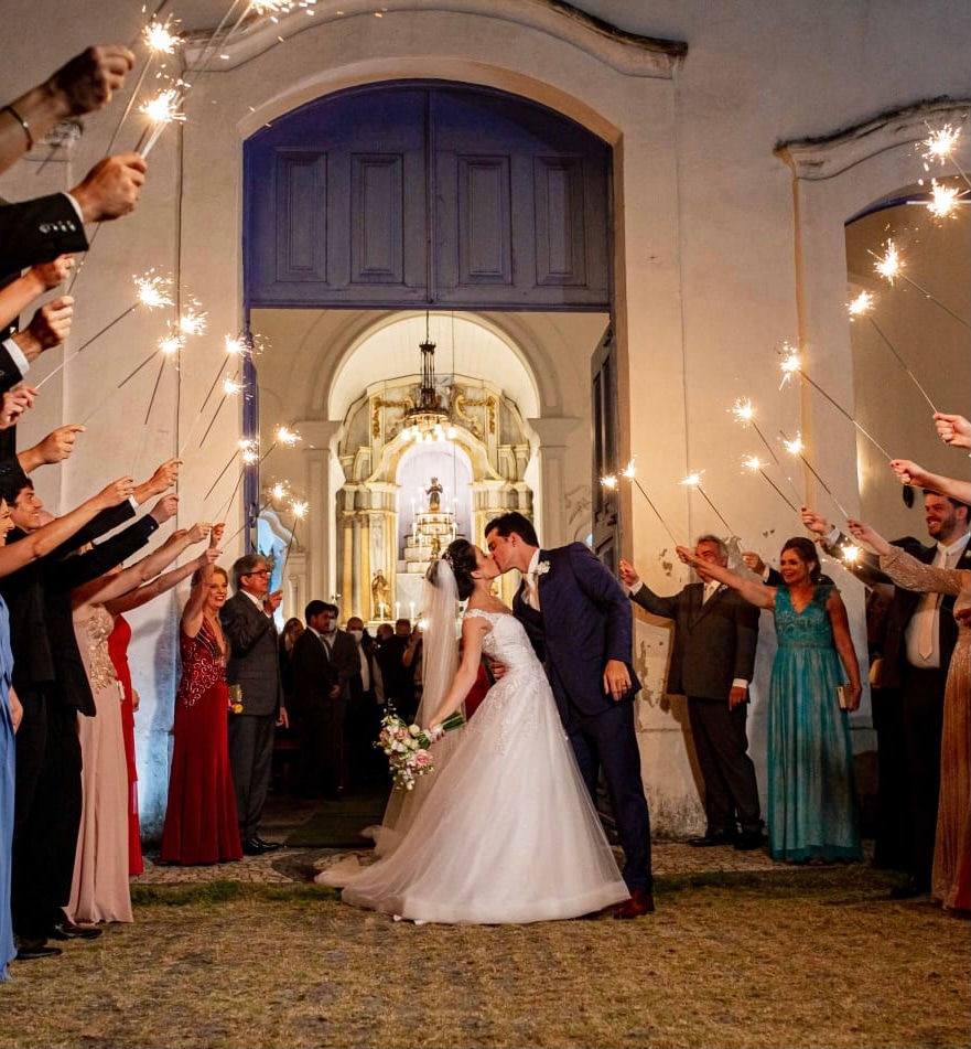 Casal se beijando na saída da igreja sob o brilho de sparklers segurados pelos convidados em ambiente noturno.