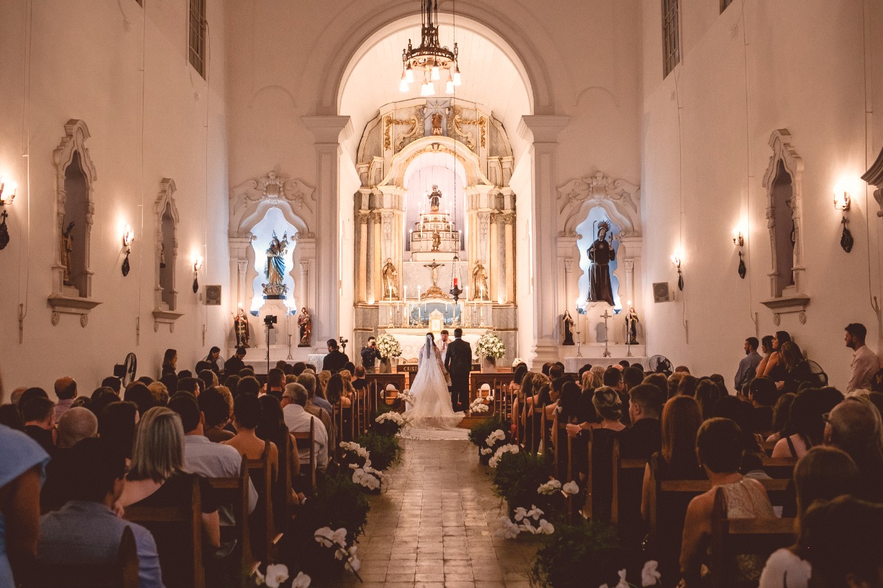 Vista do fundo da Igreja de São Gonçalo lotada durante cerimônia de casamento, destacando a arquitetura barroca e os noivos no altar.