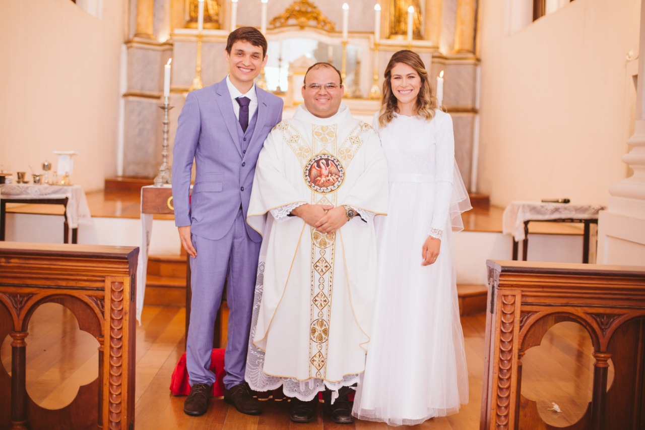 Noivo de terno azul claro e noiva de vestido de manga longa posando ao lado do padre no centro da igreja.