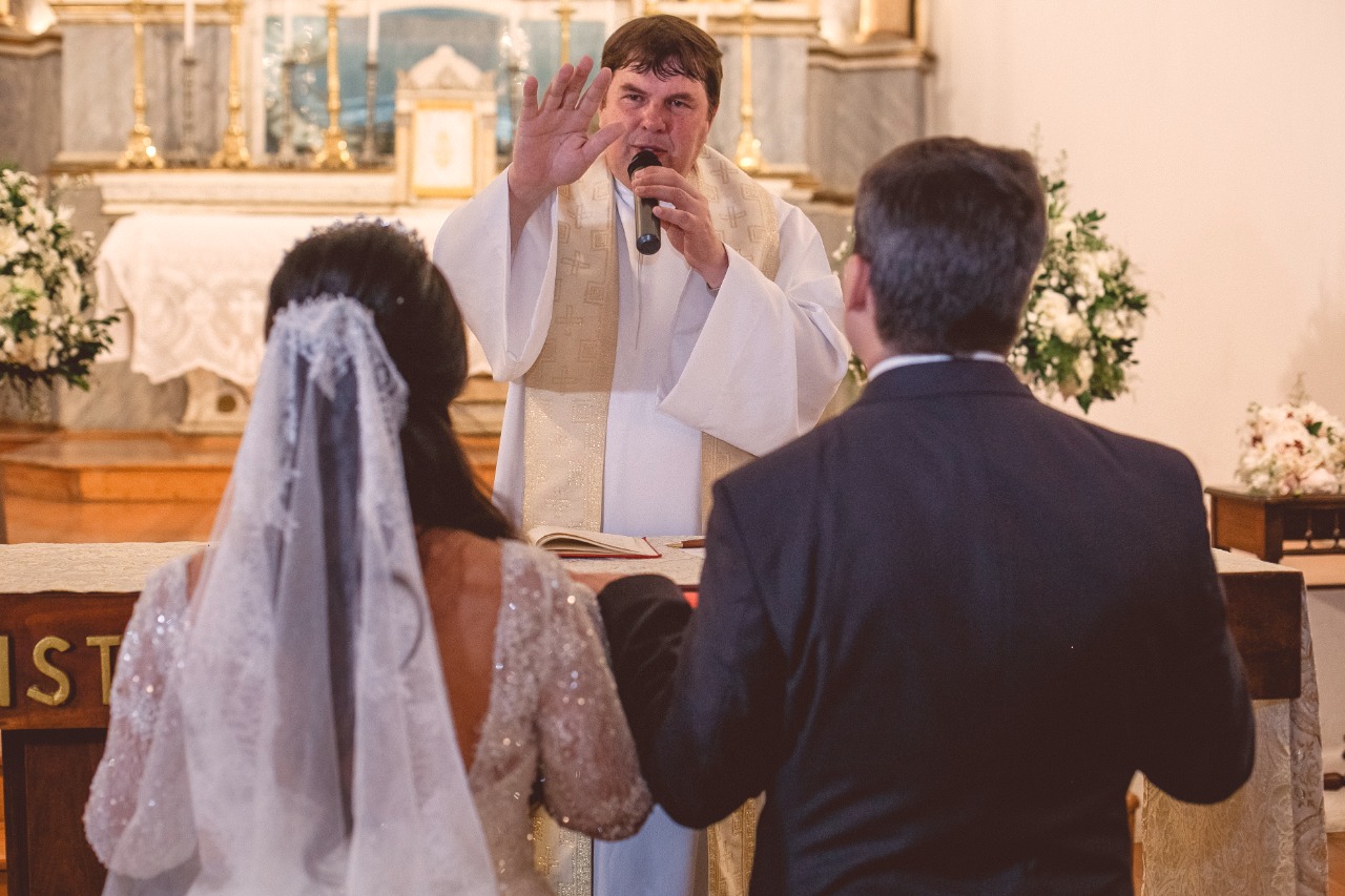 Padre abençoando casal de noivos de costas durante cerimônia de casamento na igreja.