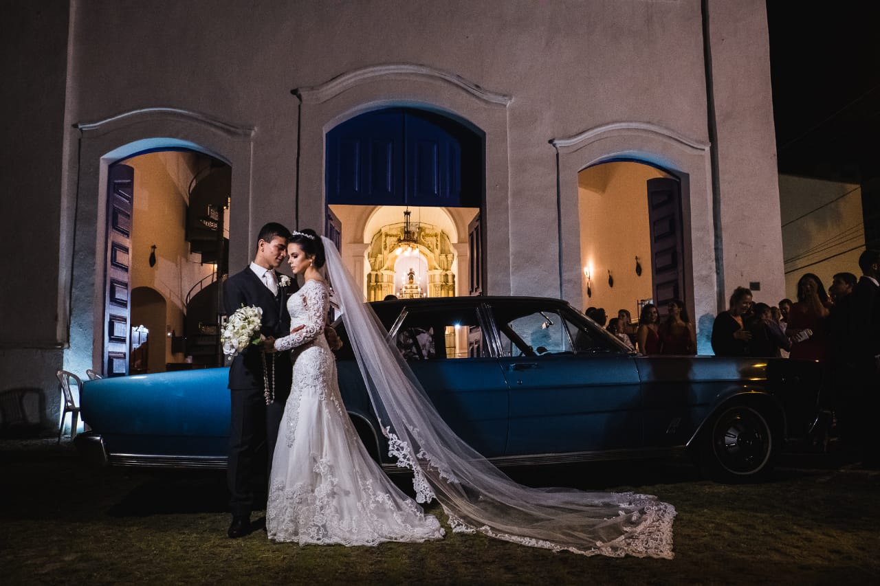 Noivos posando à noite em frente à fachada da igreja ao lado de um carro clássico azul antigo.