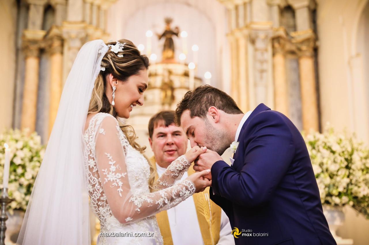 Close do noivo beijando a mão da noiva com ternura durante a cerimônia, com o padre ao fundo presenciando o momento.